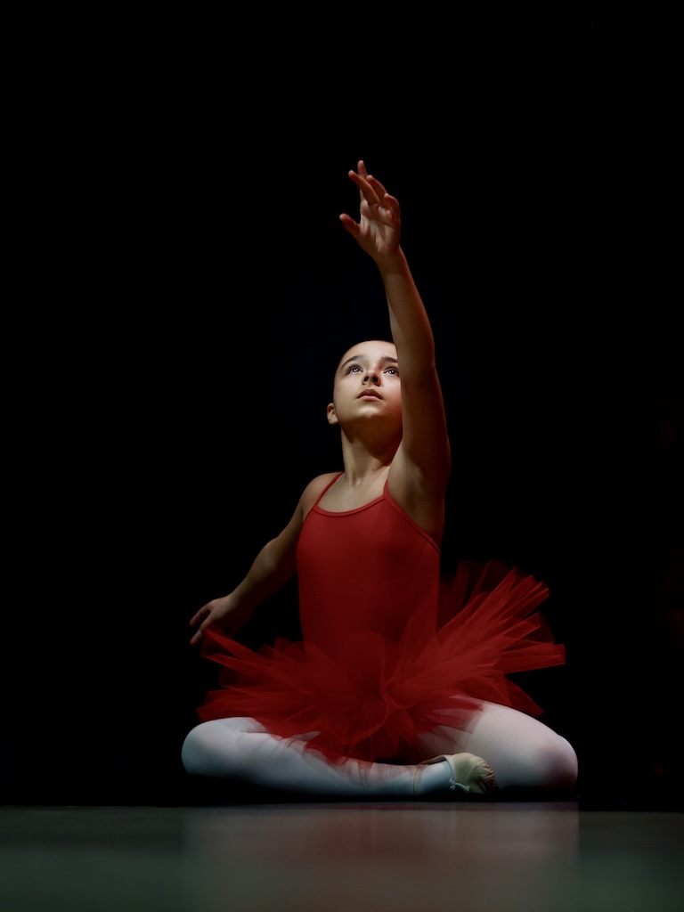 A young ballerina dressed in red reaching up.